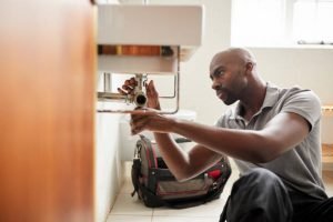 Young black male plumber sitting on the floor fixing a bathroom sink, seen from doorway